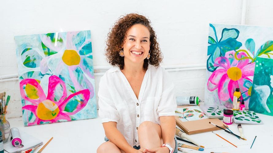 Ros Gervay smiling, sitting among her bright flower paintings